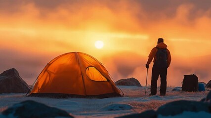 A lone backpacker stands beside a glowing orange tent watching sunrise over frosty mountains. Concept of fearless exploration and mindful wilderness solitude.