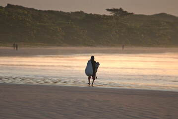 Surfer silhouette walking on the beach sand early morning before sunrise. Light reflections on sky and water. Itaguare beach, Bertioga, Brazil