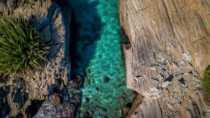 Aerial view of a clear turquoise water channel bordered by rugged, textured rocks, with lush green vegetation peeking through. Bright sunlight creates glimmers on the water's surface, enhancing the tr