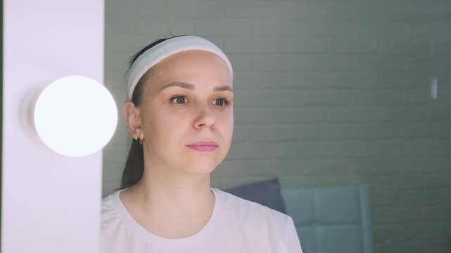 A woman in a white shirt and a headband lifts weights while looking at herself in a vanity mirror. The mirror has a row of lights around the frame.