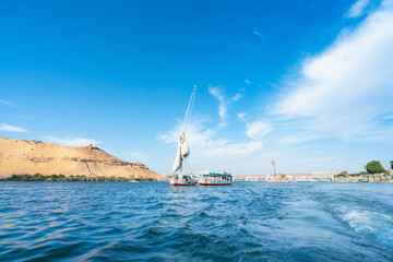 Aswan High Dam, Egypt: A felucca glides on the Nile River in spring, with sandy hills and boats along the shore under a blue sky.