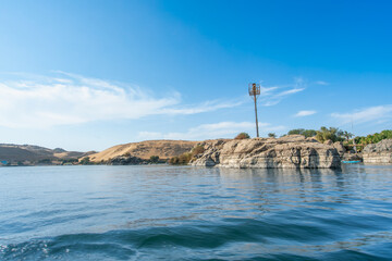 Aswan High Dam, Egypt: A serene Nile River view in spring, with rocky shores, greenery, and sandy hills under a clear blue sky.