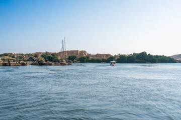 Aswan High Dam, Egypt: A scenic Nile River cruise in spring, with a boat near a rocky shore and distant hills under a clear blue sky.
