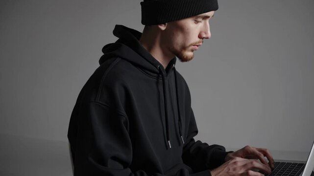 Man in black hoodie and beanie typing on laptop, representing hacker or cybersecurity professional in neutral setting

