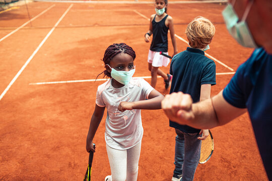 Kids wearing masks doing elbow bump with coach on clay tennis court during pandemic
