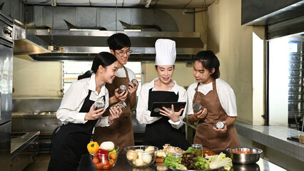 A group of women in aprons are looking at a tablet while standing in a kitchen. They seem to be discussing something related to the food they are preparing.