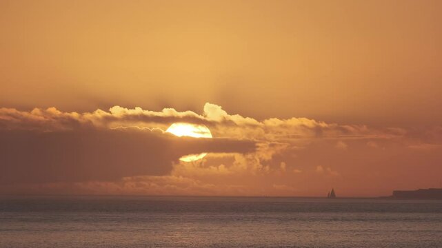 Golden sunlight softly illuminating sailboat gliding across calm atlantic waters during sunset, clouds partially veiling radiant sun near portuguese coastline