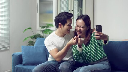 Young asian couple creating korean heart sign while capturing playful selfie moment on smartphone, expressing romantic connection and digital lifestyle at home