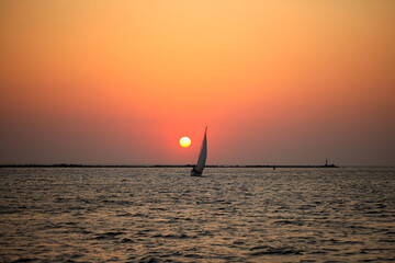 Sailboat at sunset over calm sea horizon Silhouette of a sailboat gliding across the ocean at sunset with a glowing orange sky