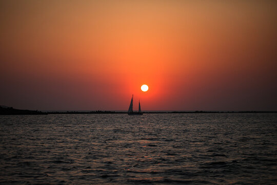 Sailboat at sunset over calm sea horizon Silhouette of a sailboat gliding across the ocean at sunset with a glowing orange sky