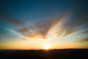 Sun sets behind rolling hills, casting warm, golden glow across sky. Wispy clouds, tinged with hues of orange and yellow, stretch across deepening blue sky. Serene and expansive landscape.