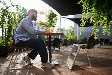 Man works on laptop at outdoor round table, with glass of iced coffee. Solar panel nearby, emphasizing sustainable, modern eco-friendly workspace that combines technology and sustainability.