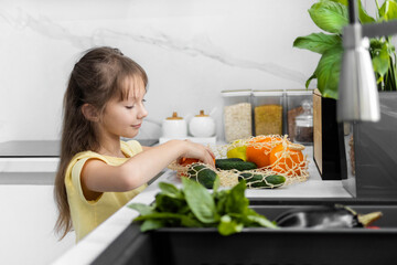 A little girl takes out organic vegetables and fruits from a mesh bag in the modern kitchen. Grocery shopping. Zero Waste. Eco-Friendly Shopping. The concept of healthy lifestyle.