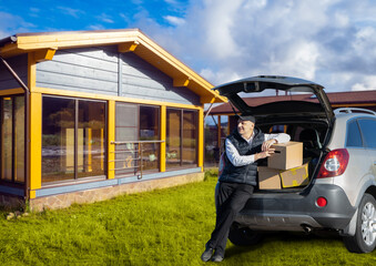 Man sitting on car trunk with cardboard boxes near modern country house, unloading or moving in on...