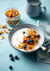Delicious bowl of yogurt topped with crunchy granola and ripe blueberries. A jar of granola and a jug of milk are in the background.