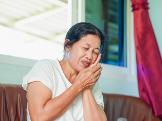 Asian senior woman holding the side of her mouth with her hand that has a sore tooth. Molar pain. Lifestyle, health care and medicine. Elderly people having a toothache. Self care at home. 