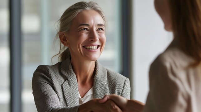 A mid aged business woman is smiling warmly as she shakes hands with a colleague in a modern office. The atmosphere is friendly and professional, fostering collaboration and teamwork