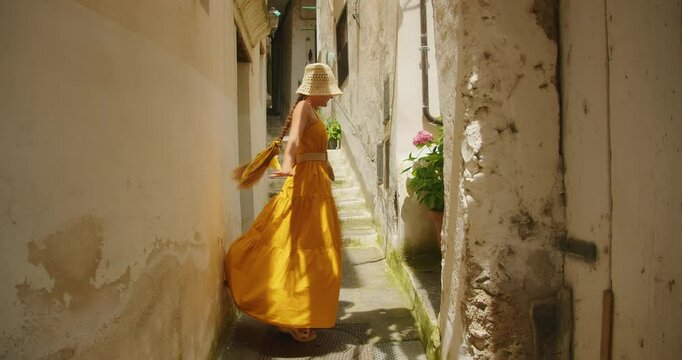 Woman in a flowing yellow dress and straw hat walks through a narrow sunlit alley in an old Mediterranean town on Amalfi coast in Italy