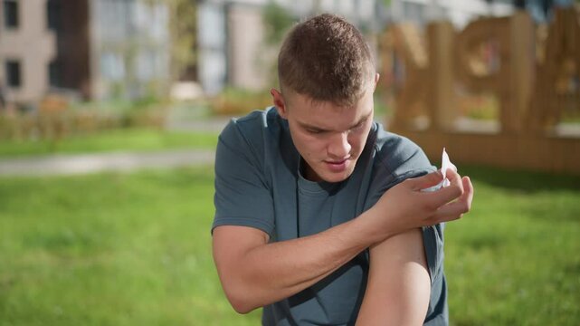 young boy seated in sunlight lifts shirt sleeve and carefully applies nicotine patch to upper arm while surrounded by soft blur of green outdoor environment suggesting self-care and wellness action