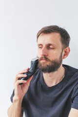 Young man aged 30 35 grooming beard under chin with electric trimmer, looking up, wearing black...