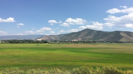 Handheld camera out of the window of a train passing through Heber Valley Utah.  Lush green meadow and Wasatch Mountain Range, puffy clouds and blue sky. Passing through Heber Utah on a train.