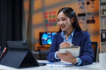 Fototapeta premium Smiling businesswoman working late at night in office using tablet and taking notes on notebook