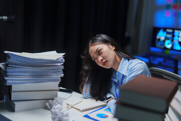 Asian office worker looking stressed while working late at night with a big pile of paperwork and books on her desk