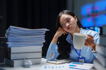Tired employee drinking coffee and looking at a large pile of paperwork while working late at night in the office