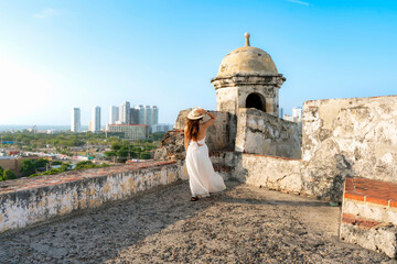 Obraz premium Taken at sunset in Cartagena, Colombia, a Unrecognized young Latina woman in a white dress and hat walks through San Felipe Castle, enjoying the breeze and panoramic city view.
