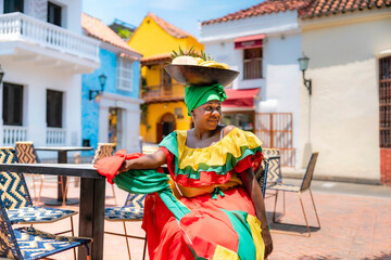 Afro-Latina Palenquera woman in colorful red, green, and yellow outfit sitting with fruit bowl on...