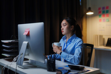 Young Asian businesswoman holding a coffee cup while working late at night in the office using...
