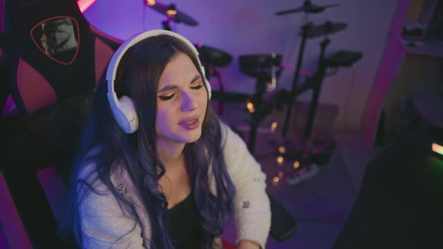 Overhead view of music artist wearing headphones covering face with hands in frustration, seated in gaming chair surrounded by studio equipment, with illuminated drum kit and mood lighting