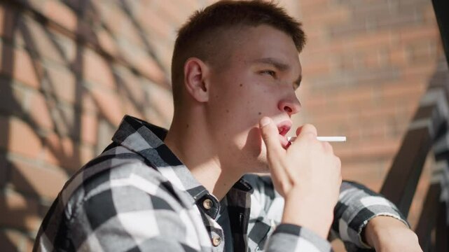 side view of man resting on iron railing smoking cigarette gazing thoughtfully into distant wearing check flannel shirt and jeans warm sunlight casts geometric brick and staircase
