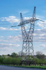 Duży stalowy słup elektryczny na tle wiosennego nieba i kwitnącego sadu,wyróżnia się na tle czystego, błękitnego nieba z rozproszonymi chmurami. Na pierwszym planie widoczny jest kwitnący sad .