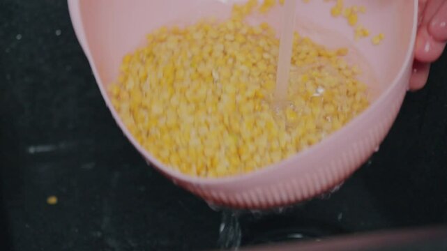 A person rinses yellow split peas in a pink colander, allowing water to wash away impurities.