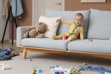 Naughty baby boy with toys sitting on sofa in messy room
