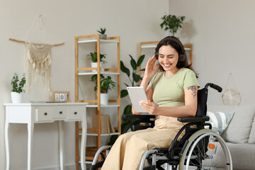 Beautiful young woman in wheelchair with headphones using tablet computer at home