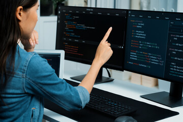 Young Asian girl of IT developer pointing online information on pc with coding program data application on website project, wearing jeans shirt. surrounded by safety programing two screens. Stratagem.