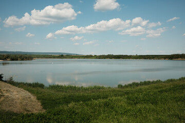 Scenic view of Pavlovac Lake in Serbia on a sunny day. Calm water reflects the sky and clouds, surrounded by green fields and distant hills. Peaceful nature and countryside atmosphere.