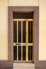 Elegant golden metal entrance door with dark glass, ornate grille, and built-in mail slot, set in a yellow wall with red stone frame. Front view. Urban architecture detail.