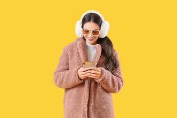 Stylish young woman in winter clothes using mobile phone on yellow background