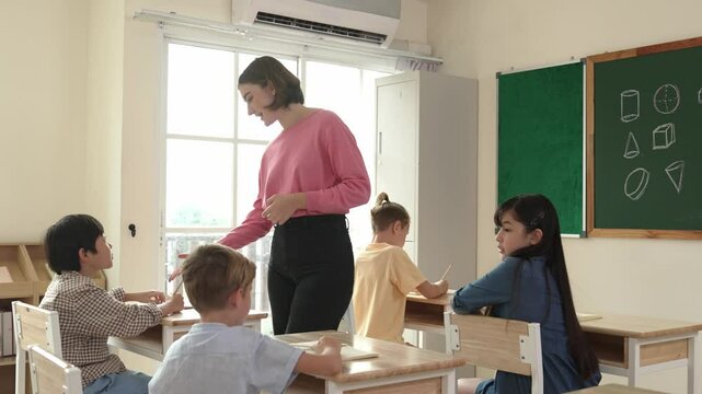 Caucasian teacher walking and check student homework at classroom while diverse children doing classwork at classroom. Diverse smart student lecturing and taking a note at elementary school. Pedagogy.