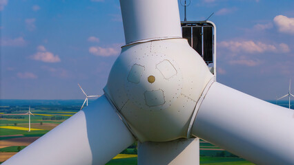 Close up on a wind turbine on a sunny day, surrounded by golden rapeseed fields and lush greenery,...