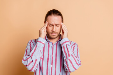 Handsome man with beard and brown hair in a striped shirt holding head in pain against a beige background