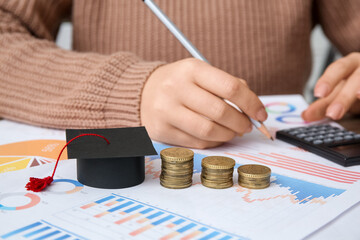 Woman with graduation hat and coins writing at table, closeup. Student loan concept