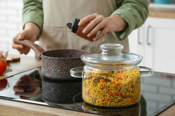 Cooking pot with pilaf on stove against woman in kitchen, closeup