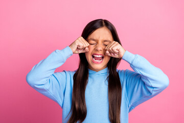 Girl in blue sweatshirt rubbing her eyes against pink background with expressive gesture showcasing...