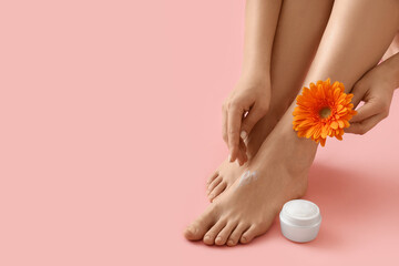 Young woman with flower applying body cream on her legs against pink background
