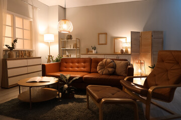 Interior of living room with brown sofa, armchair and glowing lamps in evening