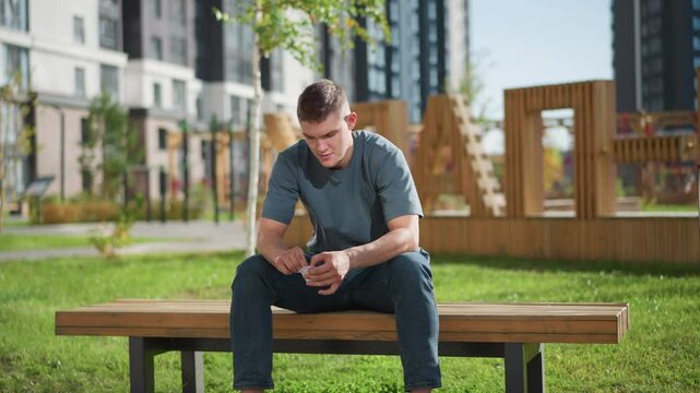 young man sitting on bench outdoors opening nicotine patch pack with paper backing falling behind him on grass next to residential buildings under daylight promoting quit smoking